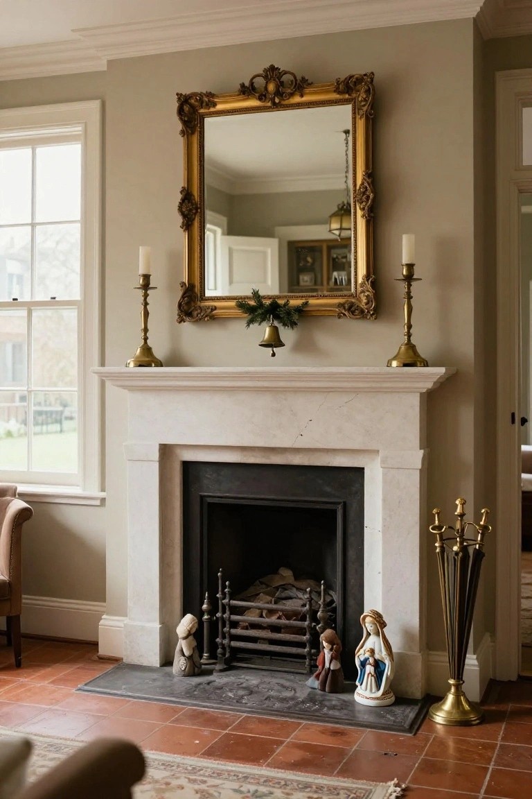 A fireplace mantel in a pale green-walled room with a large ornate gold-framed mirror centered above the firebox, flanked by brass candlesticks holding white candles, Christmas greenery on the mirror, small ceramic figurines on the hearth, and a brass fire tool stand nearby.