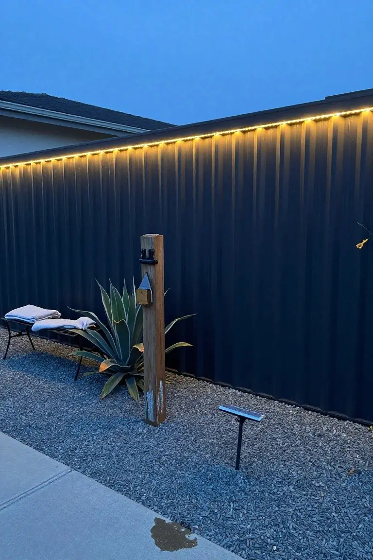 Tall dark corrugated metal fence with continuous yellow LED strip lights along the top edge at dusk, wooden post with attachments and birdhouse, agave plant, metal bench with towels, solar stake light, gravel ground, and concrete path.