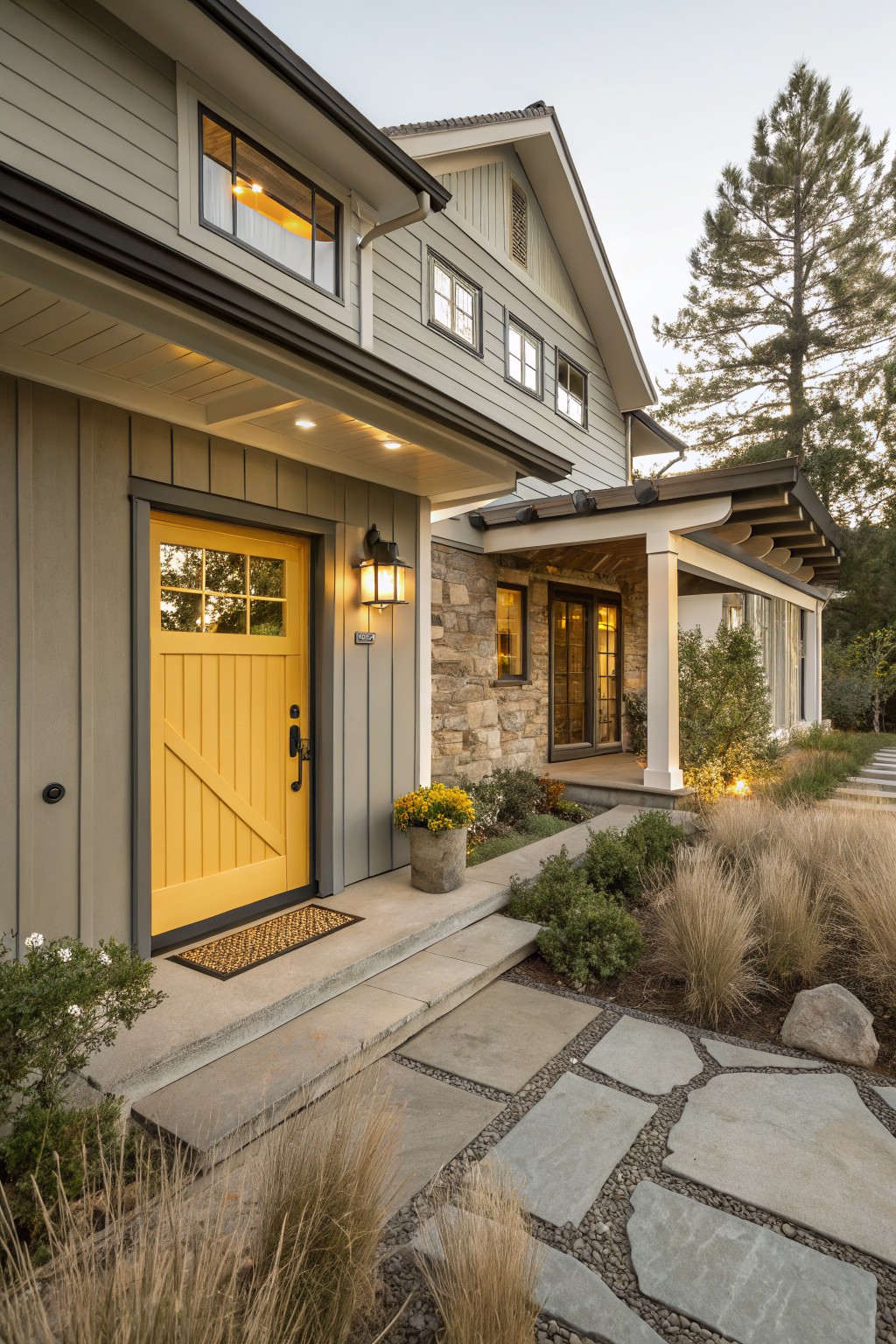Gray board-and-batten sided house exterior with yellow paneled front door, stone accent wall, covered porch, lantern light, stone steps, pathway, and grasses.