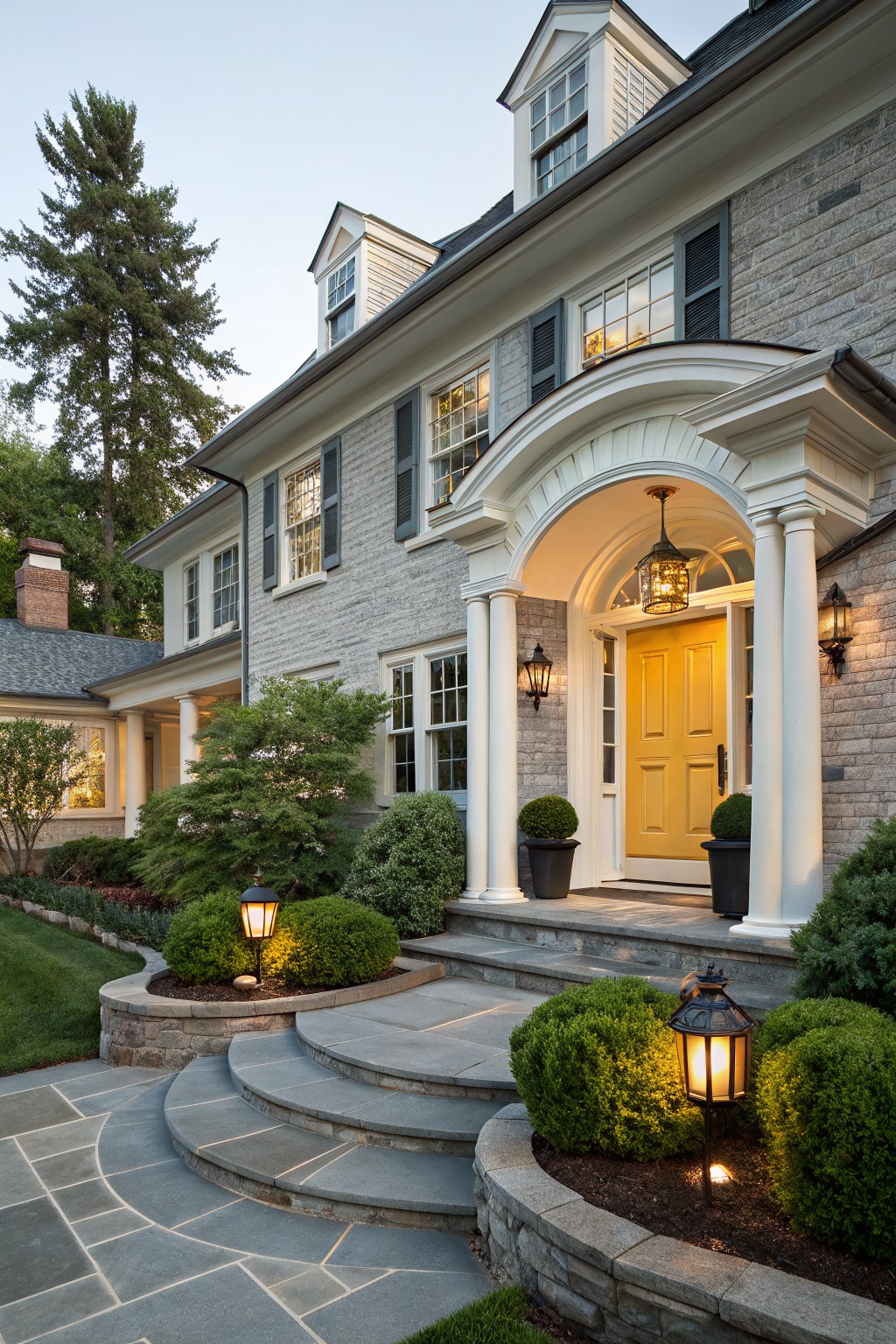 Gray stone two-story house exterior featuring a yellow front door under a white arched portico with columns, flanked by lanterns, stone steps, shrubs, and a tall pine tree nearby.