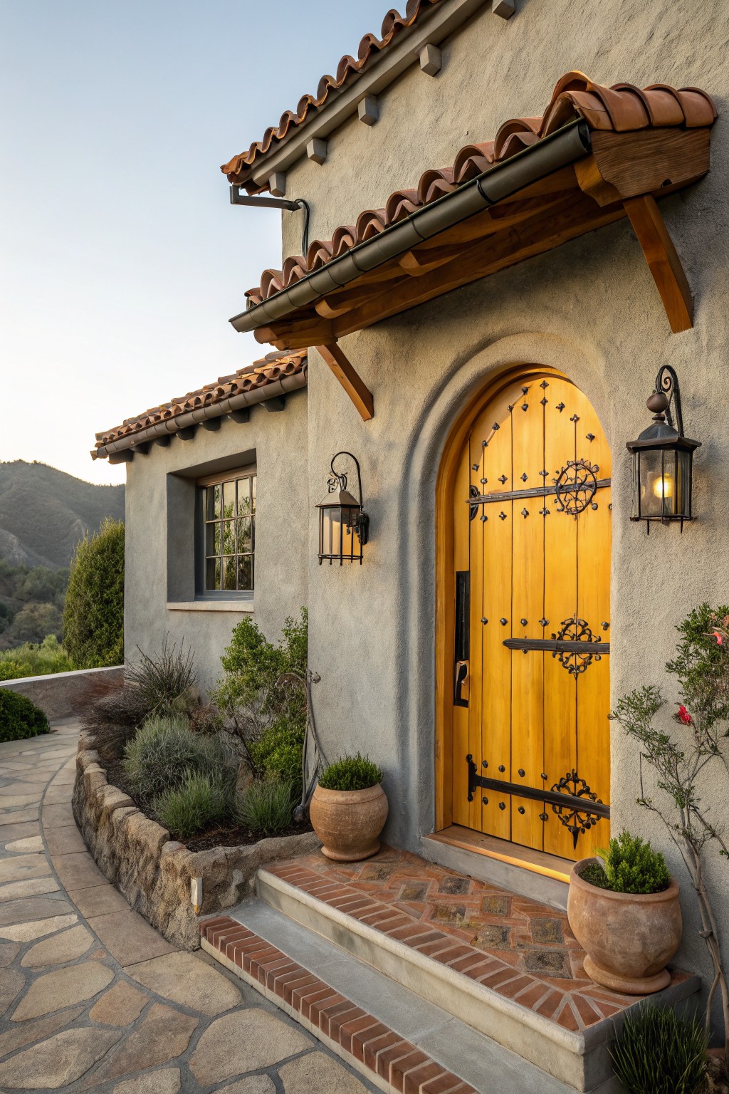 Gray stucco house exterior with red tile roof, featuring a tall yellow arched wooden door with black wrought iron hardware and hinges, flanked by black lanterns, stone steps, and potted plants along a curved pathway.