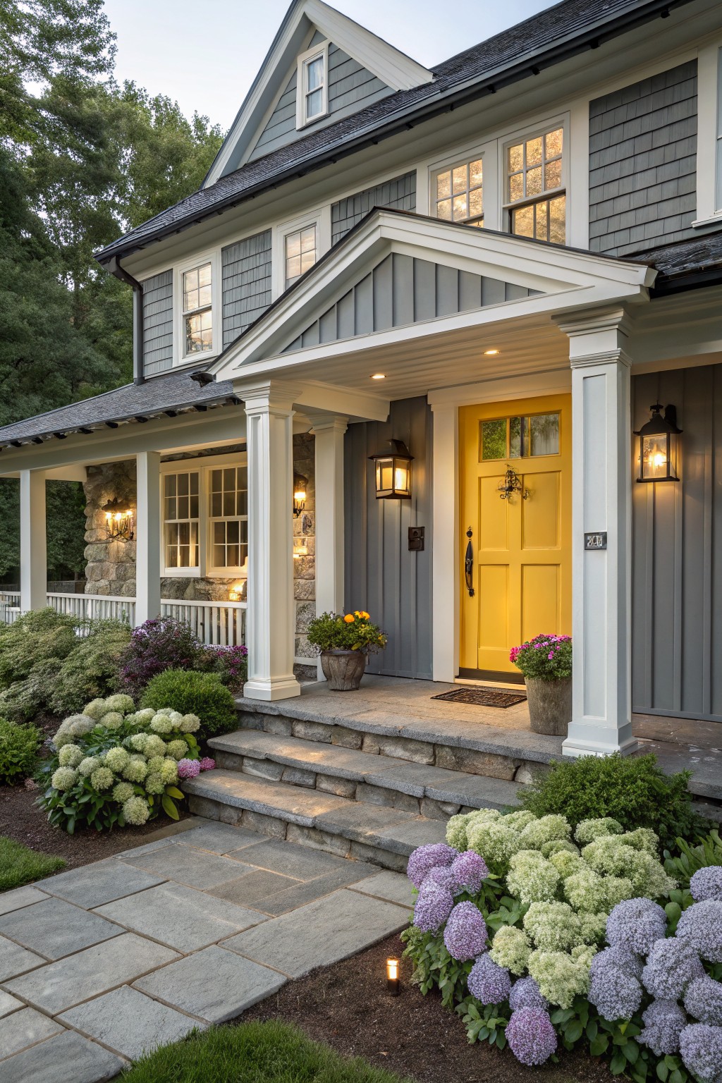 Gray shingle house exterior featuring a bright yellow front door with glass panels, white porch columns, stone steps, lanterns, and hydrangea plantings along the flagstone walkway.