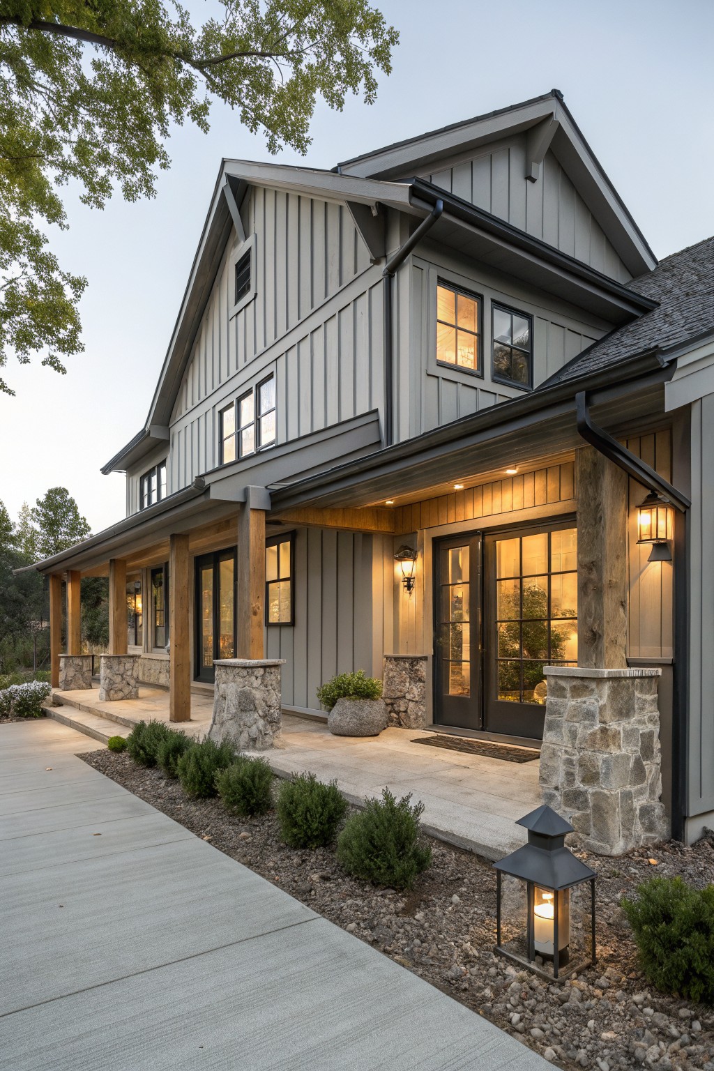 Gray board-and-batten sided house exterior with covered porch on stone pillars and wooden beams, large windows, lanterns, shrubs, and concrete walkway at dusk.