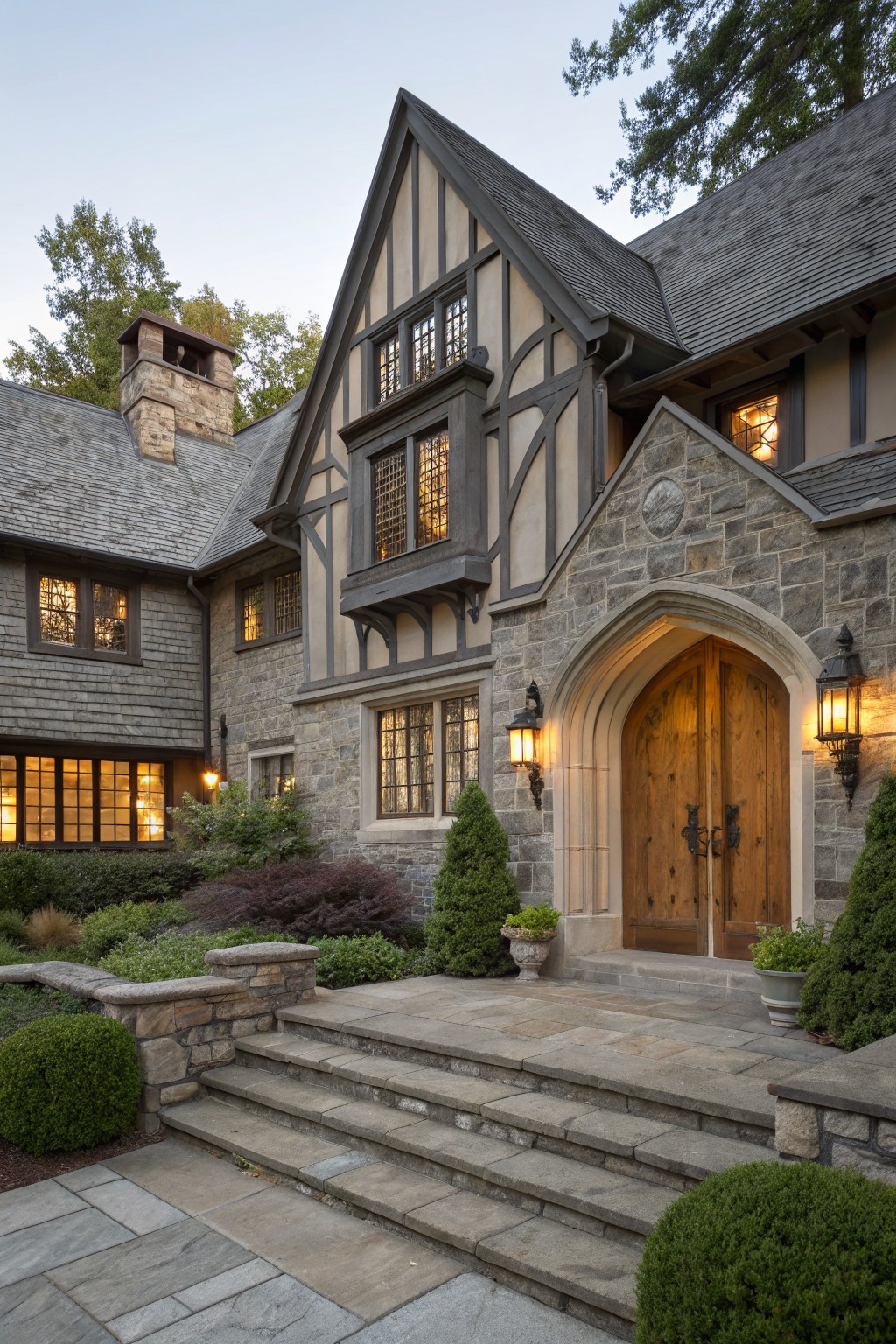 Gray Tudor-style house exterior with dark timber framing, stone-clad arched entryway featuring double wooden doors and lanterns, slate roof, and low evergreen shrubs along stone steps at dusk.