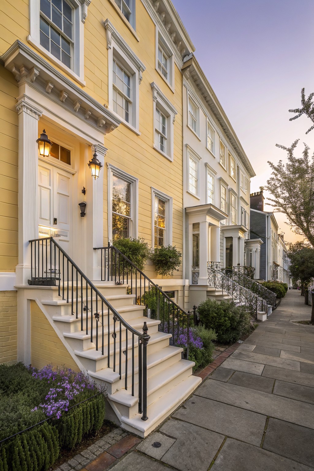 Yellow row house with white window and door trim, columns, front steps with black iron railings, lanterns by the white entry door, potted plants, flower beds, shrubs, and a sidewalk next to white and dark neighboring houses at dusk.