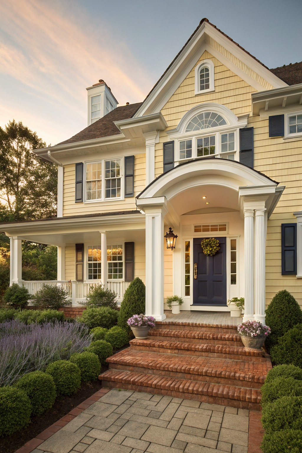 A two-story yellow clapboard house with white trim and dark blue shutters features a columned front porch with an arched entry, dark blue door, brick steps, and surrounding landscaping.