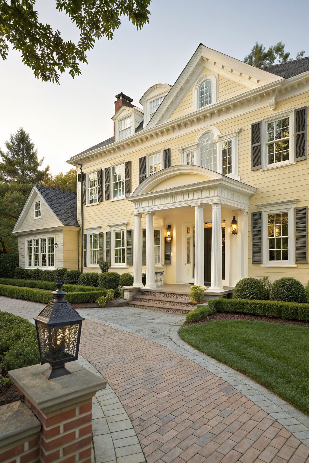 A two-story yellow clapboard house with white trim features a classical portico with white columns at the front entrance, flanked by windows with dark shutters, manicured boxwood hedges, and a curved brick pathway leading from a stone pillar lantern.