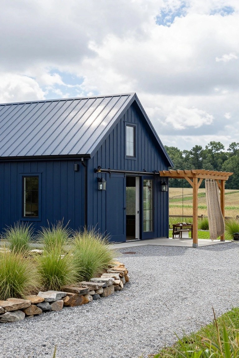 Navy blue board-and-batten sided barn-style building with metal roof, open sliding glass doors, wooden pergola with hanging fabric and chairs, gravel path, ornamental grasses, and stone edging near fields and trees under partly cloudy sky.