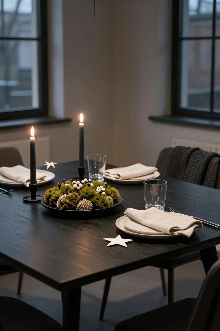 Dining table with black plate centerpiece of green moss clumps, moss balls, white berries, and pinecones, black candles in holders, white star paper cutouts, linen napkins on plates, glassware, forks, and chairs in a room with large windows and wall radiator.