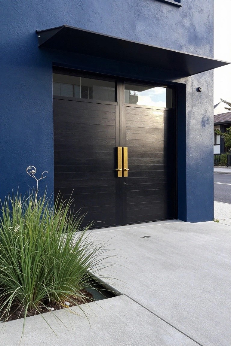 Dark blue stucco house wall with double black paneled garage doors, gold handles, metal awning overhead, and grasses planted in a narrow bed beside the concrete driveway.
