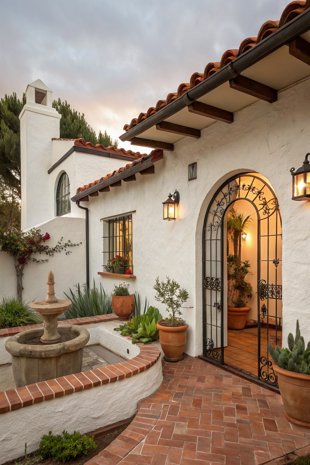 White stucco house exterior with red tile roof, arched wrought iron gate entry flanked by lanterns and potted plants, stone fountain, and brick pathway in a courtyard setting.