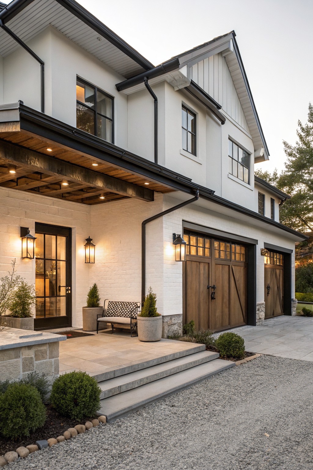 White stucco house exterior with black-trimmed windows, a covered front porch with wood beams and lanterns, double wood garage doors, stone accents, steps, potted plants, and gravel driveway at dusk.
