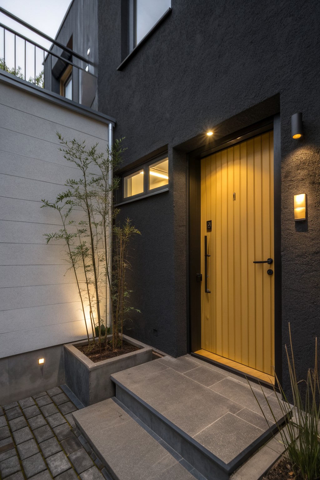 Contemporary house entrance with tall yellow vertical-plank door set in black textured walls, concrete steps, potted bamboo plants, and wall-mounted lights.