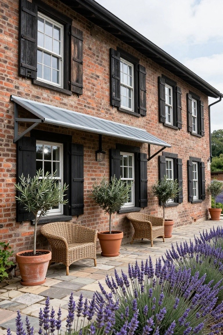 Brick house exterior featuring black wooden shutters on multipaned windows, a metal awning over one window, potted olive trees in terracotta pots, wicker armchairs on a stone patio, and a border of lavender plants.