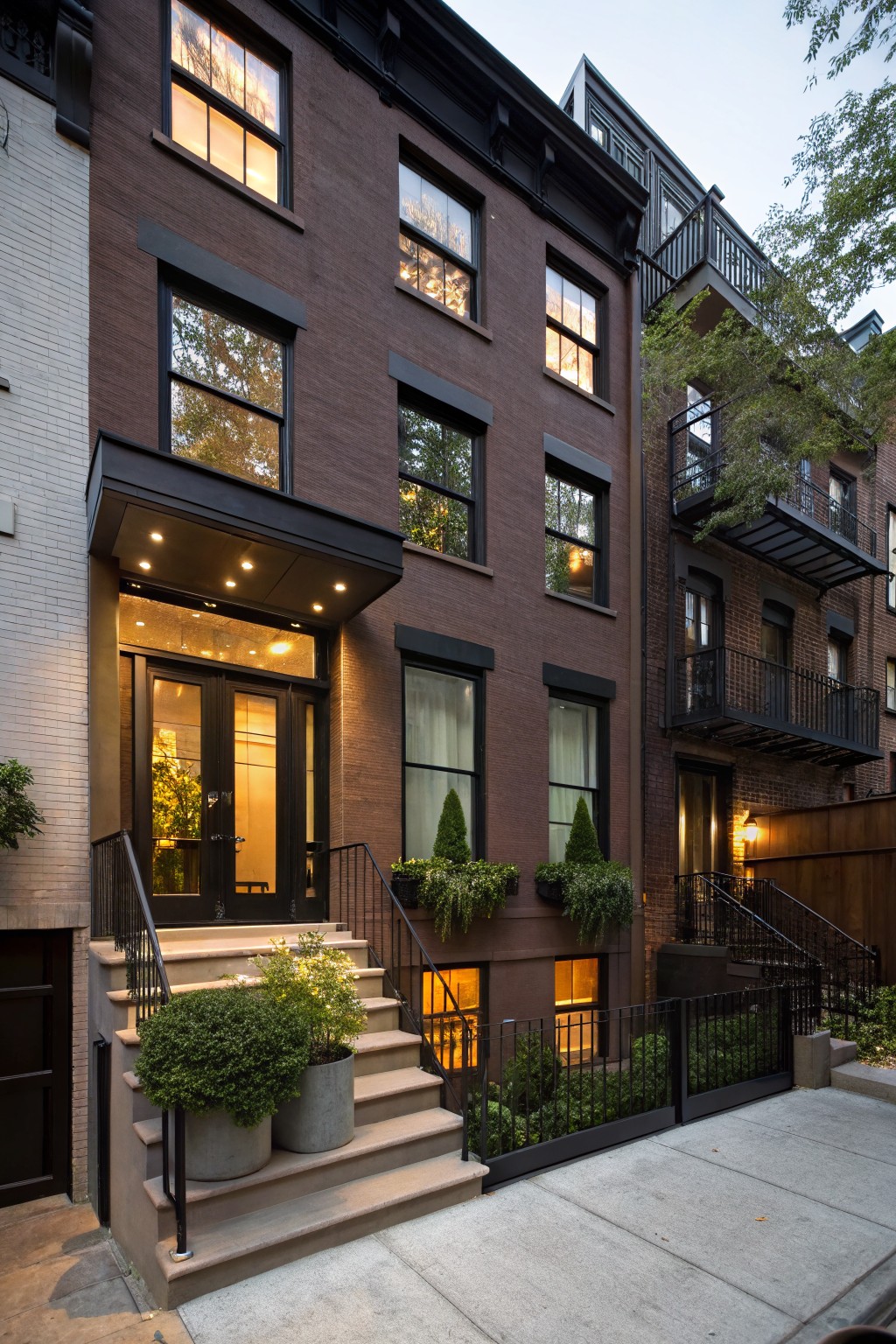 Brown brick townhouse exterior with black window frames, metal entry canopy with lights, double front doors, potted plants on steps, and adjacent buildings at dusk.