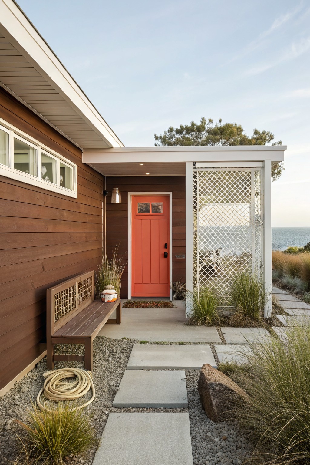 Exterior corner of a brown wood-sided house featuring a bright orange front door, wooden bench with planter, gravel ground cover, stone path, grasses, white lattice screen, and ocean view beyond.