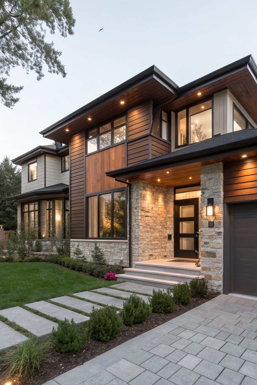 Modern two-story house exterior featuring brown wood cladding, light gray siding, stone entry pillars, large windows, black trim, and a paved driveway with landscaping at dusk.