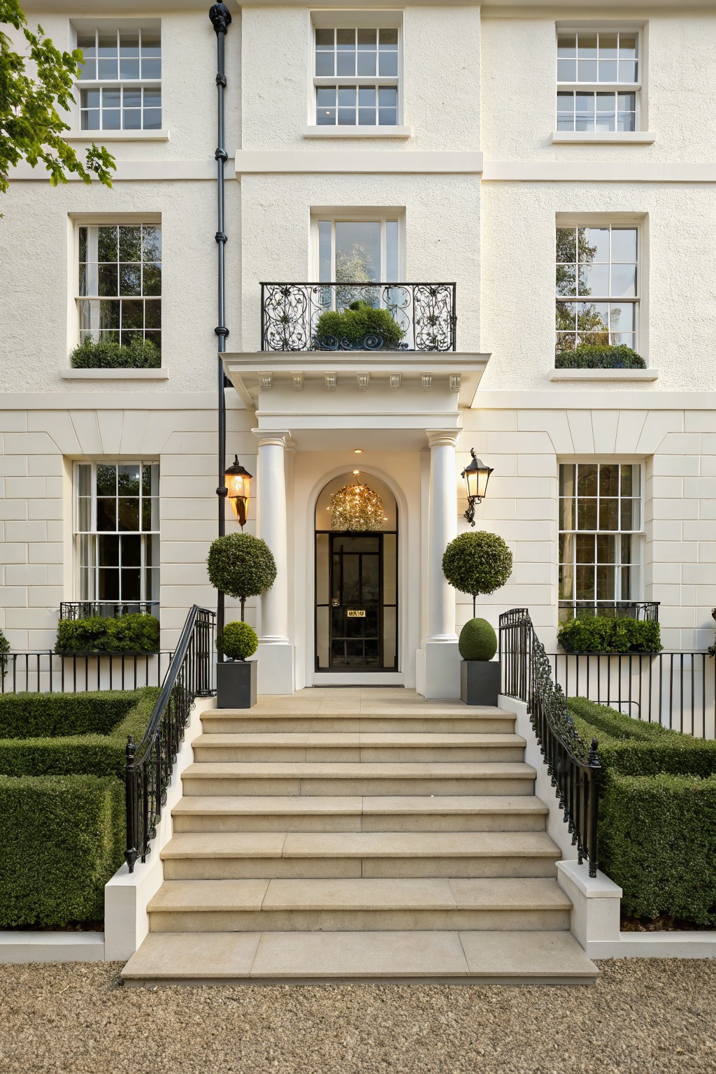 White stucco townhouse facade with stone steps leading to a black front door, flanked by spherical boxwood topiaries, low box hedges, black iron railings, and lanterns.