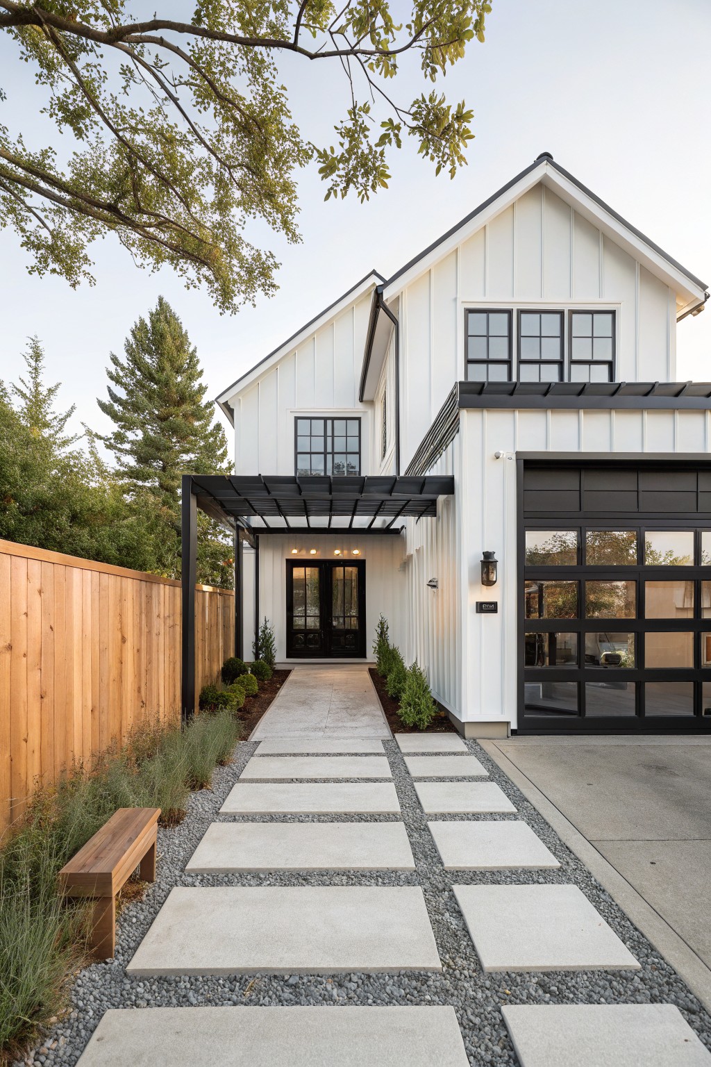White farmhouse style house with board and batten siding, black trim windows and garage door, metal pergola over entry, concrete paver path set in gravel with grasses and wooden bench leading to the door, wood fence and trees nearby.