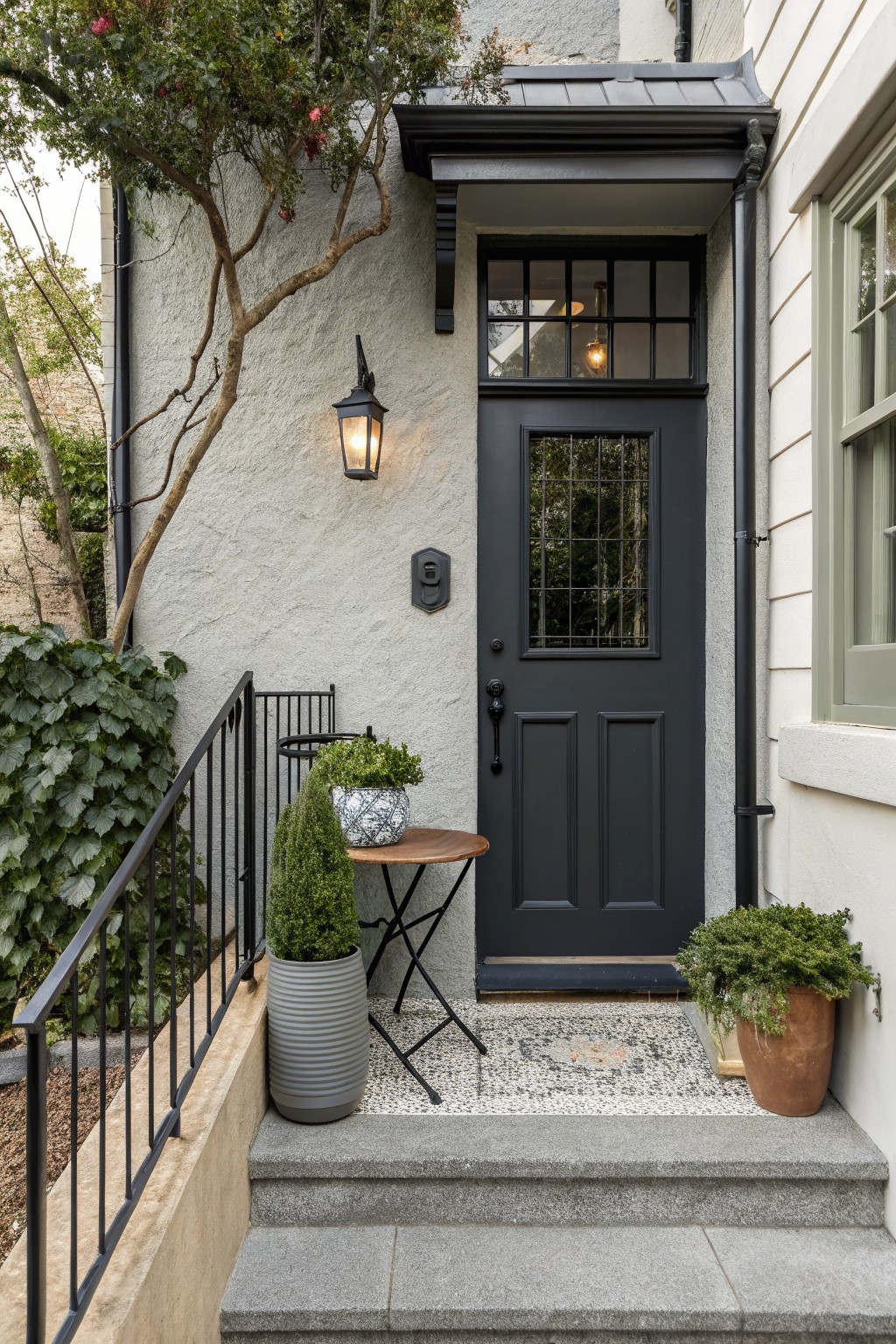 Dark paneled front door with grid glass and transom on light gray stucco house exterior, beside black lantern light, with potted plants, small round table, black metal railing, pebble landing, and stone steps.