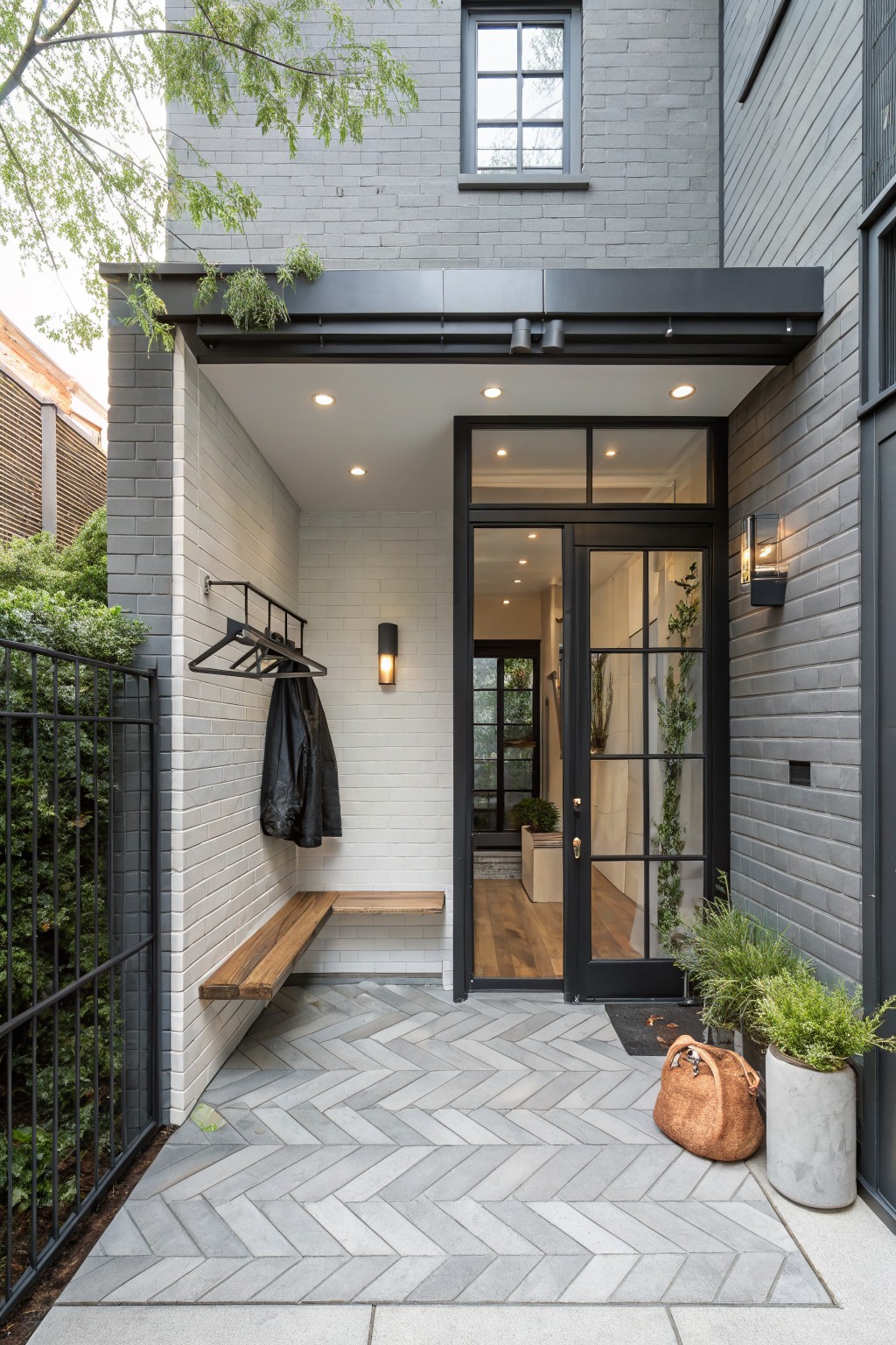 Gray brick house exterior with covered porch entry featuring herringbone-patterned gray tile floor, wooden wall bench, black metal coat rack holding jackets, black-framed glass door, potted plants, leather bag, and black metal fence with greenery.