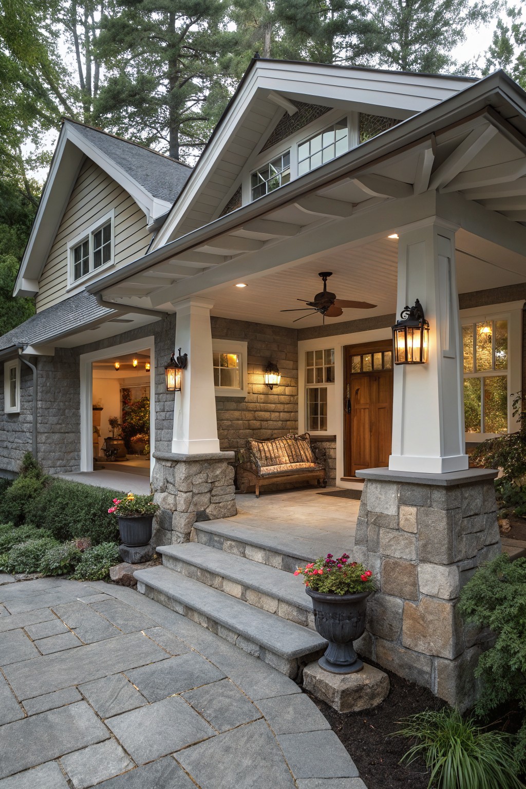 Gray Craftsman-style house exterior with open garage, front porch featuring gray stone pillars topped by white columns, wooden door with glass panels, lanterns, ceiling fan, cushioned bench, stone steps, potted flowers, and bluestone paver walkway amid shrubs and trees.