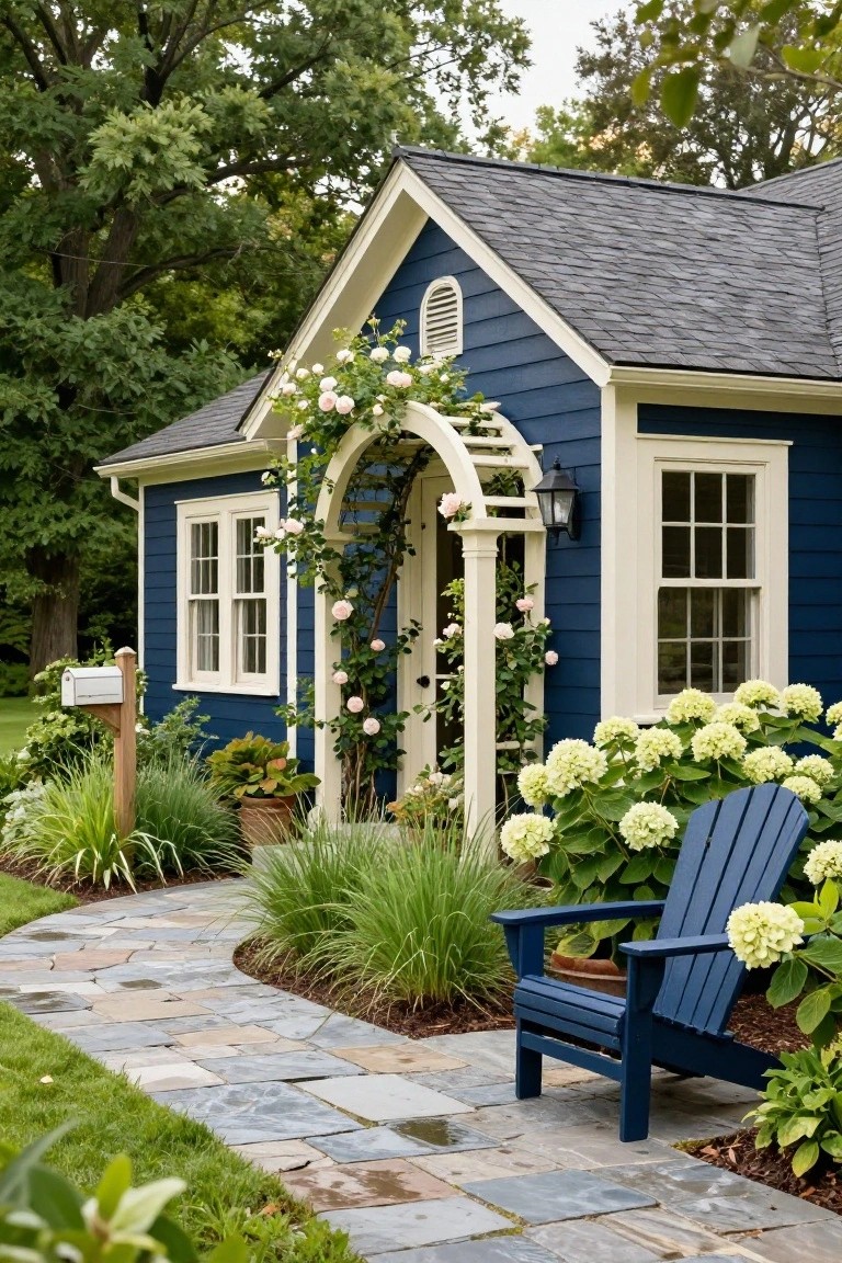 Navy blue clapboard house with white trim and gabled roof, white arched arbor over the entry covered in pink climbing roses, cream columns, blue Adirondack chair by white hydrangeas, and stone walkway.