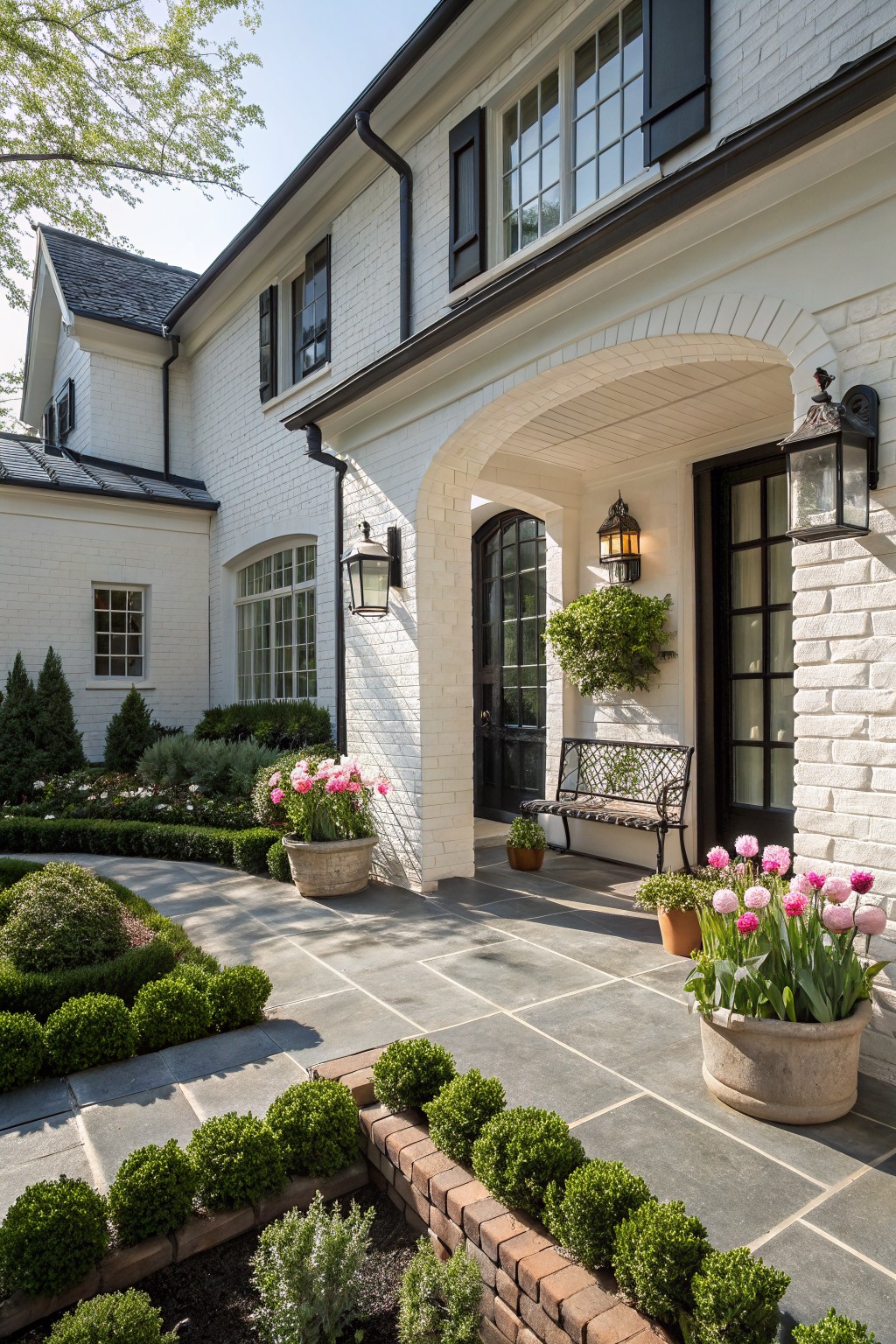 White brick house exterior with an arched portico entry featuring black double doors, wall lanterns, potted tulips, a bench, boxwood hedges, and a stone path.