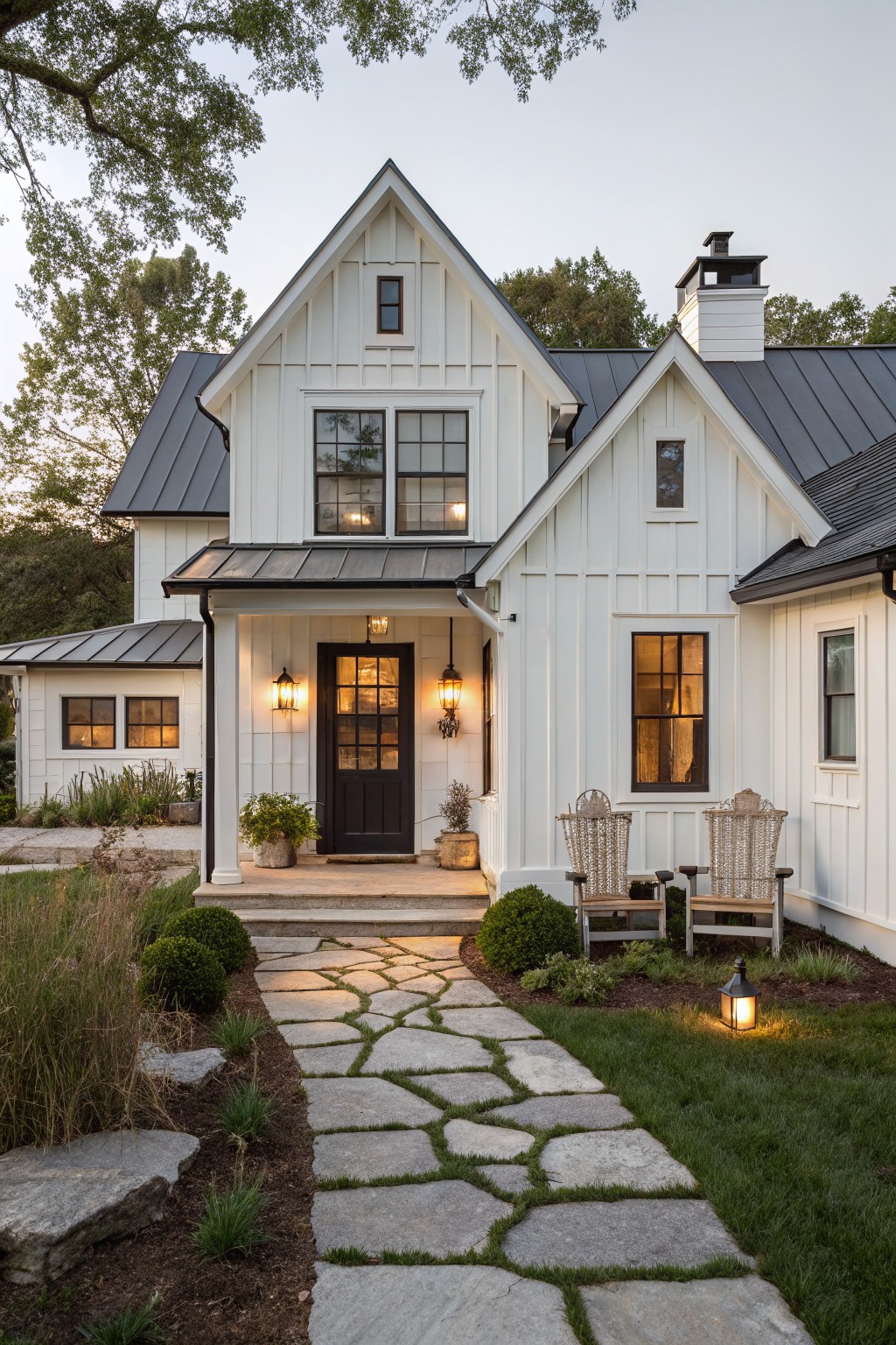 White board-and-batten farmhouse with dark metal gable roofs, covered front porch featuring black door and lanterns, stone slab pathway, potted plants, chairs, and lawn edging in evening light.