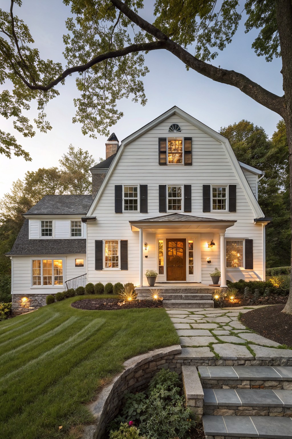 White clapboard farmhouse exterior with gambrel roof, black shutters, covered front porch featuring wood door, stone steps and pathway leading up from landscaped yard at dusk.