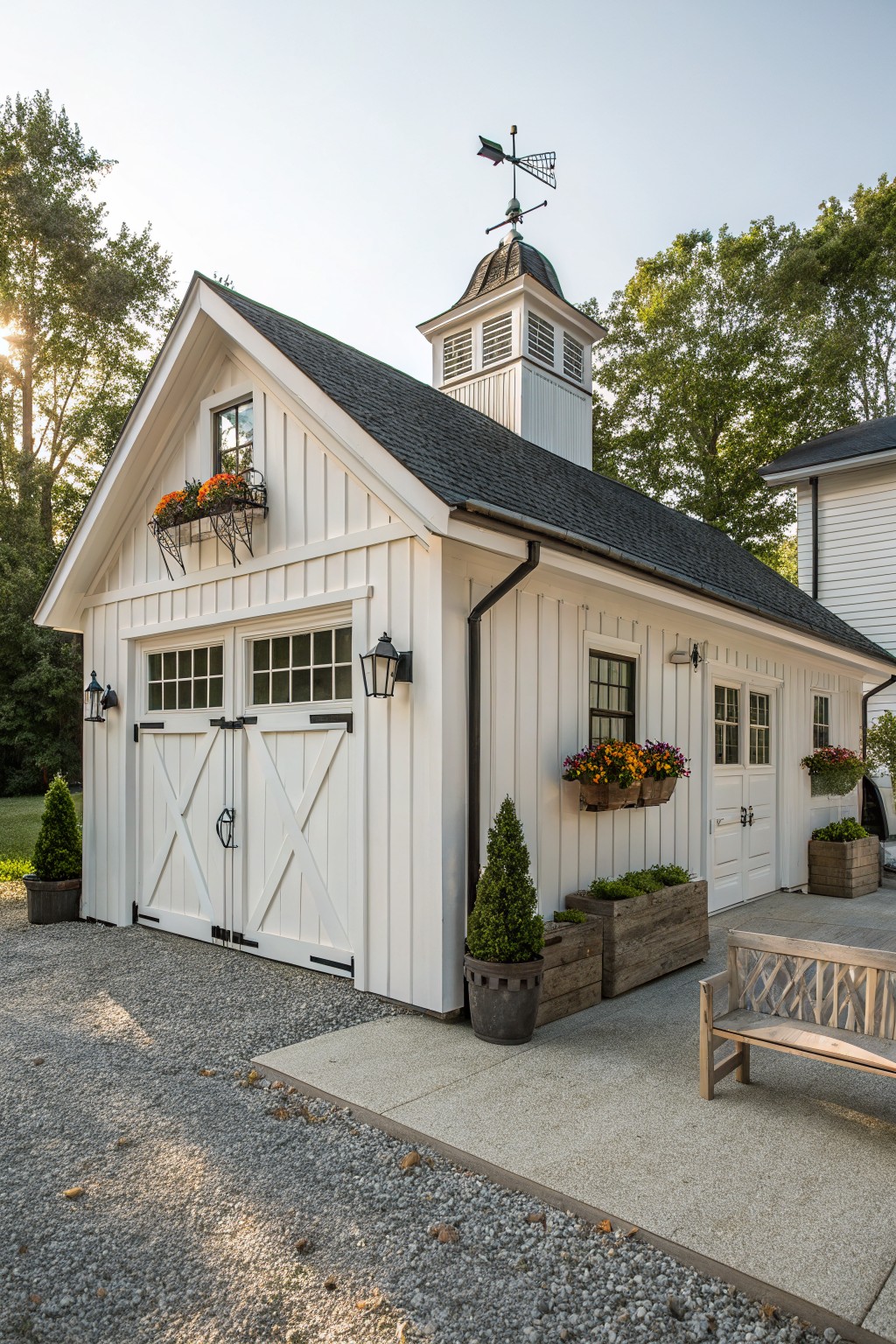 White board-and-batten detached garage with double garage doors, cupola and weather vane on peaked roof, flower boxes, wall lanterns, potted plants, gravel driveway, and trees nearby.