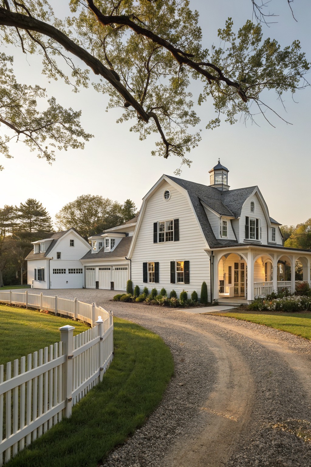White clapboard farmhouse with gambrel roof, cupola, black shutters, wraparound porch, attached garage with double doors, white picket fence, gravel driveway, and surrounding trees and landscaping at sunset.