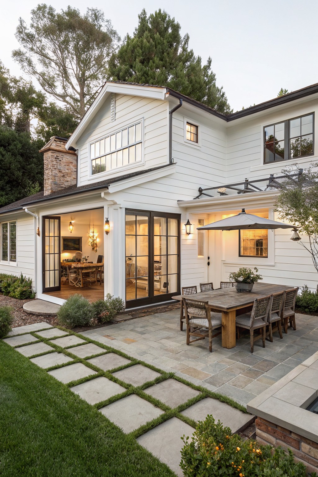 White board-and-batten farmhouse exterior with tall stone chimney, covered porch featuring open glass doors and interior table with chairs, adjacent outdoor dining table on stone paver patio, grass paths, and surrounding landscaping.