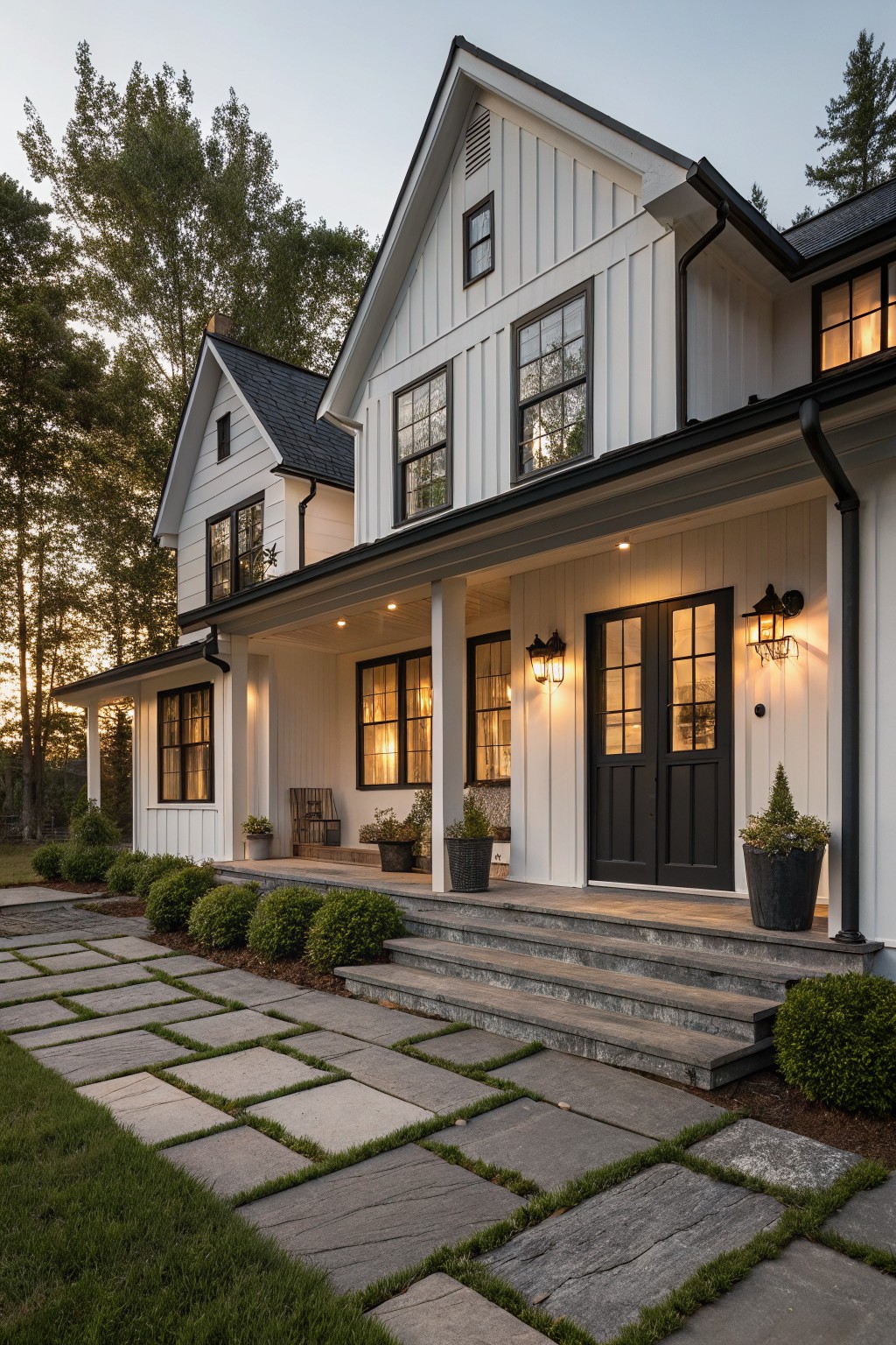 White board-and-batten farmhouse exterior with gabled roof, covered porch featuring black-framed double doors, lanterns, wooden bench, stone steps, potted plants, and bluestone pathway edged by shrubs and grass at dusk.