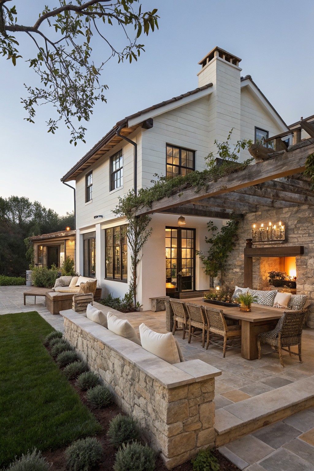 White farmhouse house side exterior with wooden pergola-covered patio, dining table and chairs with cushions, stone outdoor fireplace, low stone wall with pillows, and lawn landscaping at dusk.
