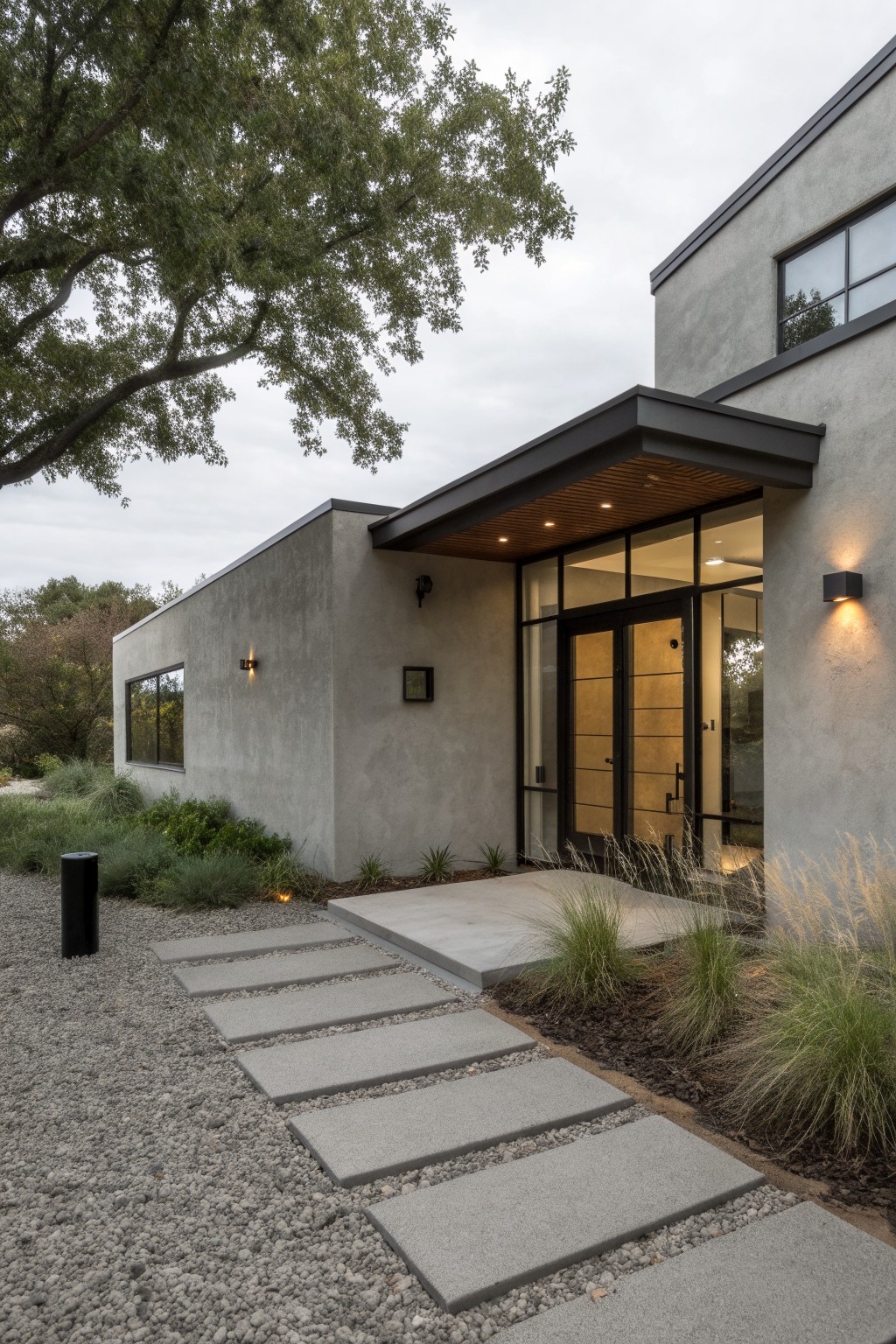 Gray stucco single-story house exterior with cantilevered dark wood entry canopy, large glass-framed double doors, wall lights, and concrete stepping stone path through gravel bordered by native grasses and plants.