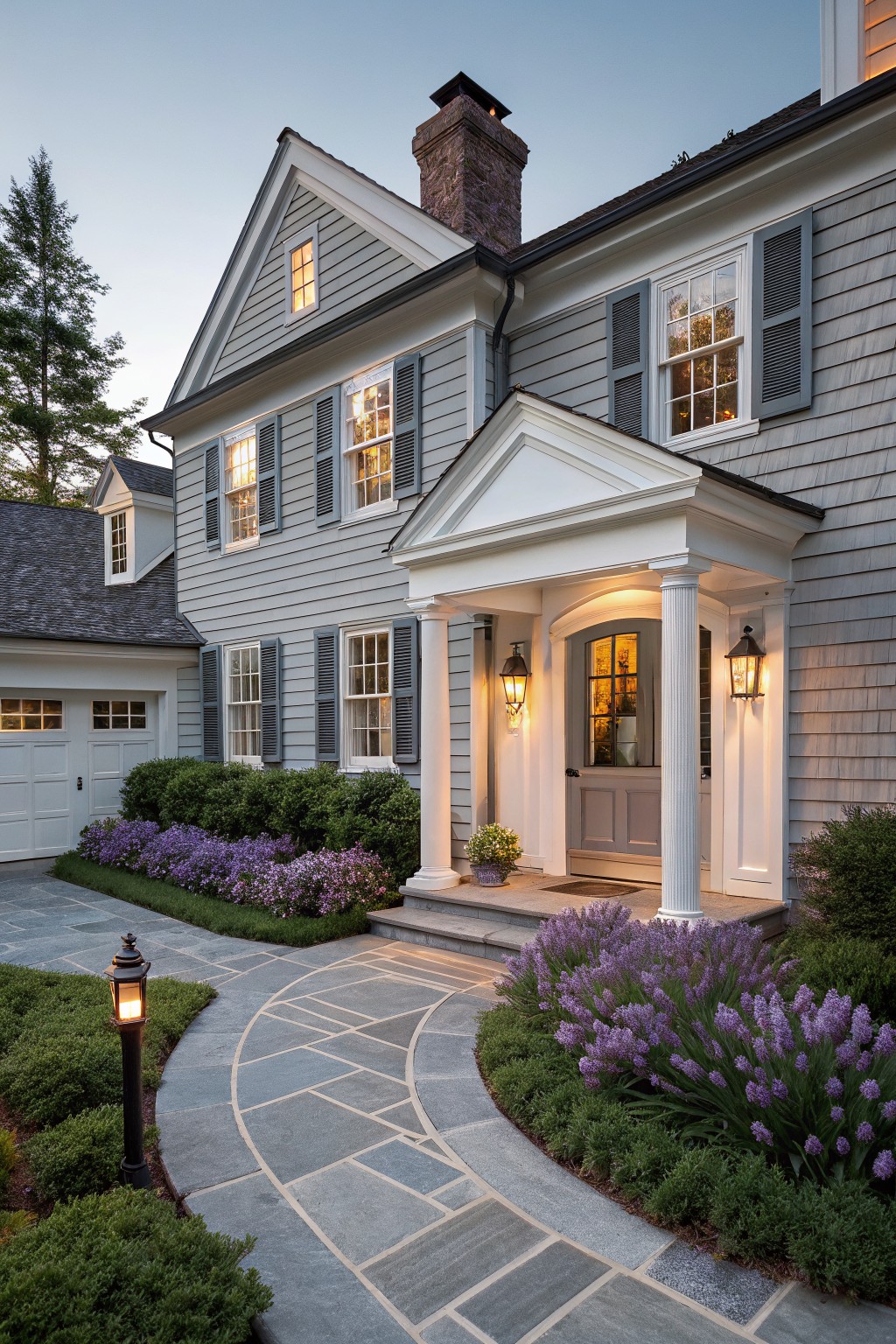 Gray shingle and clapboard house exterior featuring a white pedimented porch with fluted columns, arched door, lanterns, attached garage, and curved bluestone pathway edged with purple flowers and shrubs.
