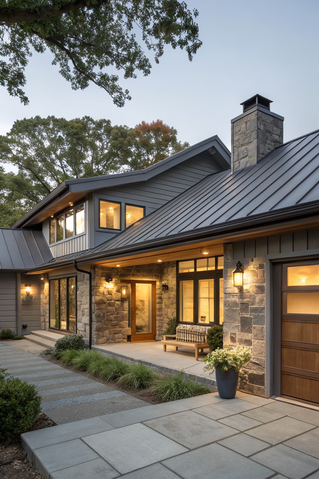 Gray ranch-style house exterior with standing-seam metal roof, stone chimney and accents, large windows, wooden entry door, garage door, covered porch, stone pathway, ornamental grasses, and potted plants in evening light.