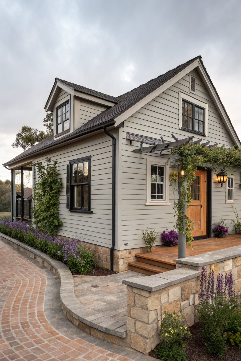 Gray clapboard ranch house exterior featuring a gabled roof, black-trimmed windows, wooden front door under a vine-covered pergola, stone base, flower beds, and curved brick pathway.