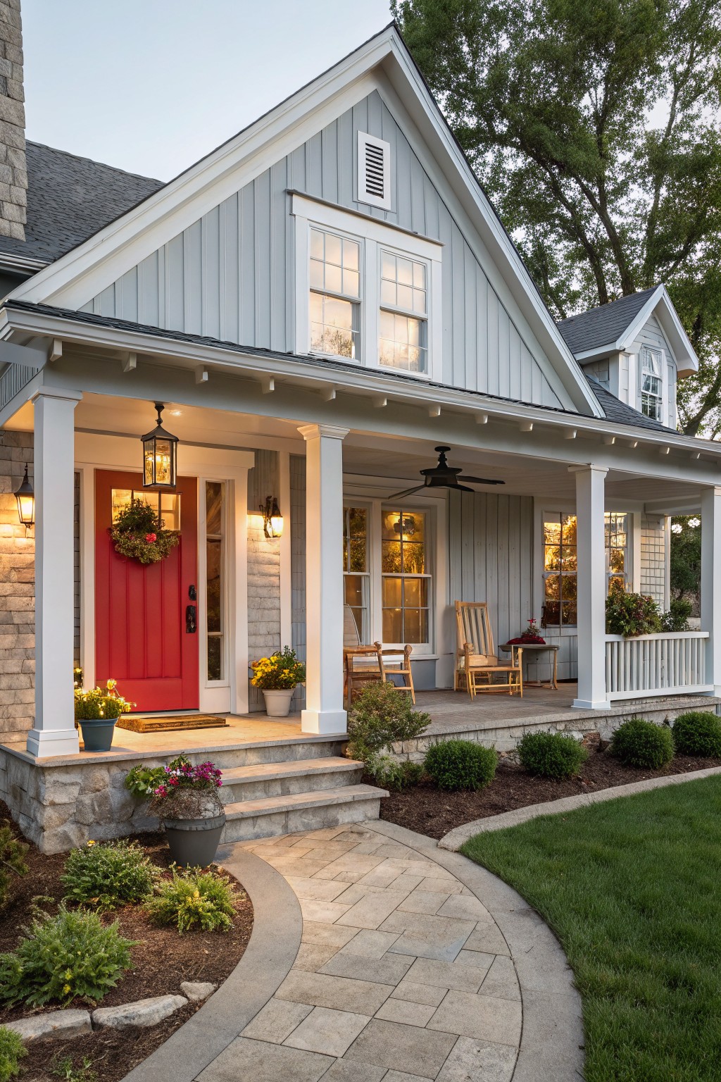 Gray clapboard-sided house exterior with gabled roof, white trim, red front door with wreath, covered porch featuring columns, ceiling fan, wooden chairs, potted plants, and curved stone pathway edged by shrubs and grass.