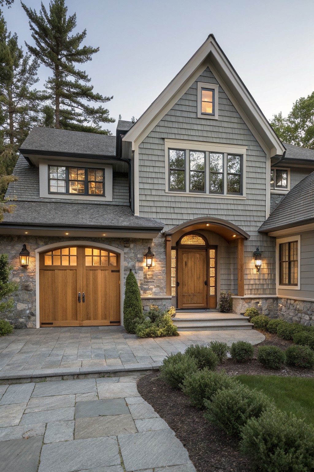 Gray shingle siding on a house exterior with stone base around wooden arched garage doors and entry door, bluestone paver driveway curving into landscaping with shrubs and trees at dusk.