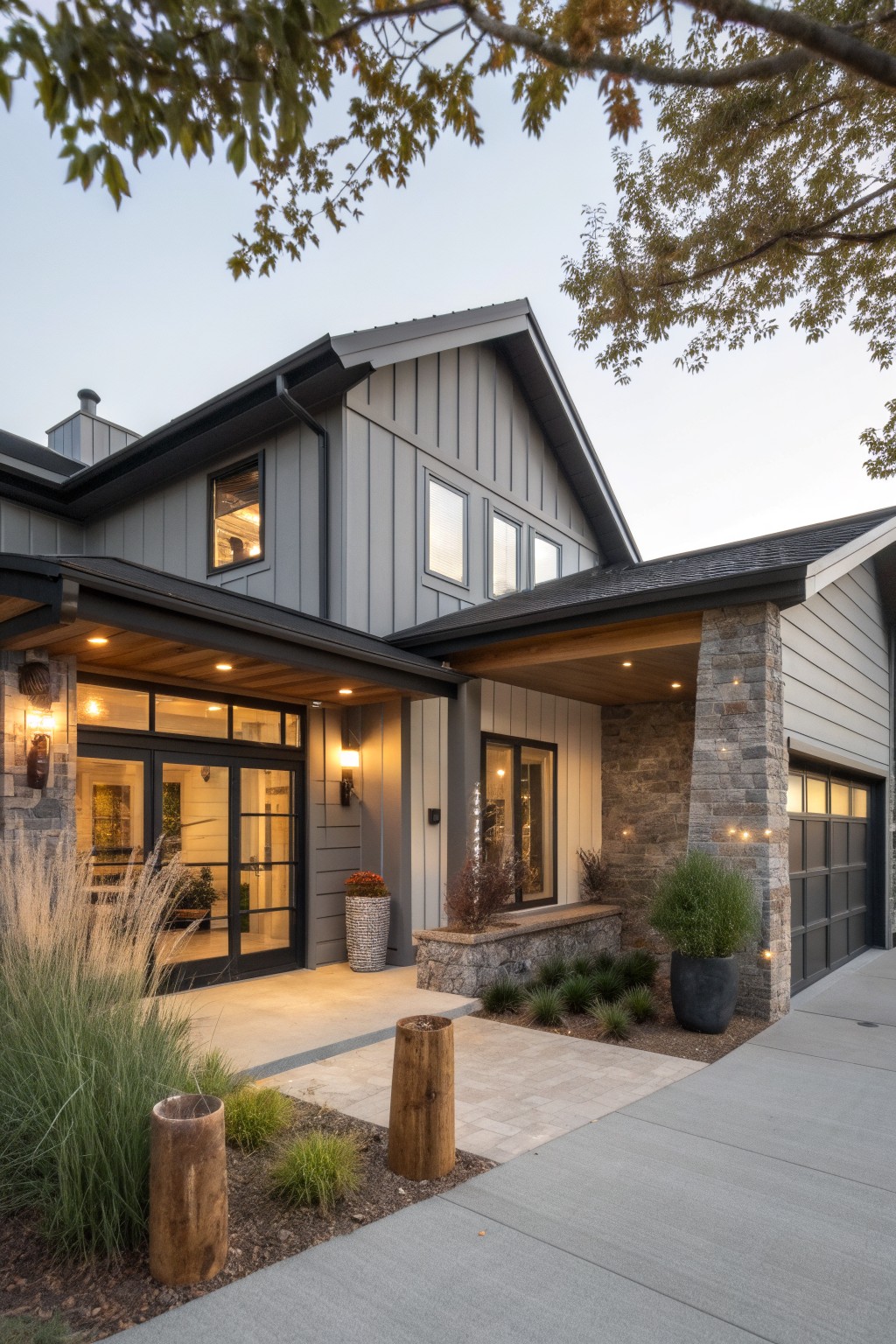 Front view of a gray board-and-batten ranch house with gabled roof, covered entry porch with wood plank ceiling and stone pillar, black-framed glass door, garage door, potted plants, and ornamental grasses along the driveway at dusk.