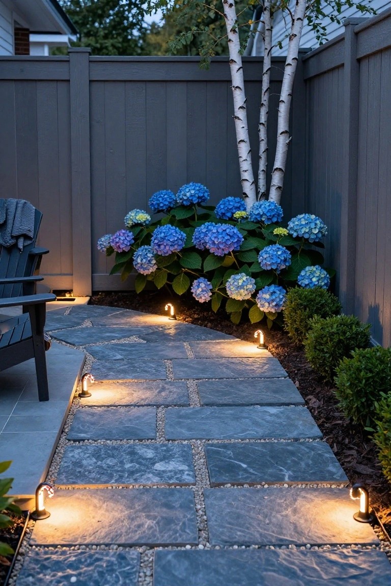 Curved stone pathway in a backyard garden lined with candy cane-shaped path lights, passing blue hydrangea bushes, birch trees, boxwood shrubs, and a dark wood Adirondack chair near a gray fence.