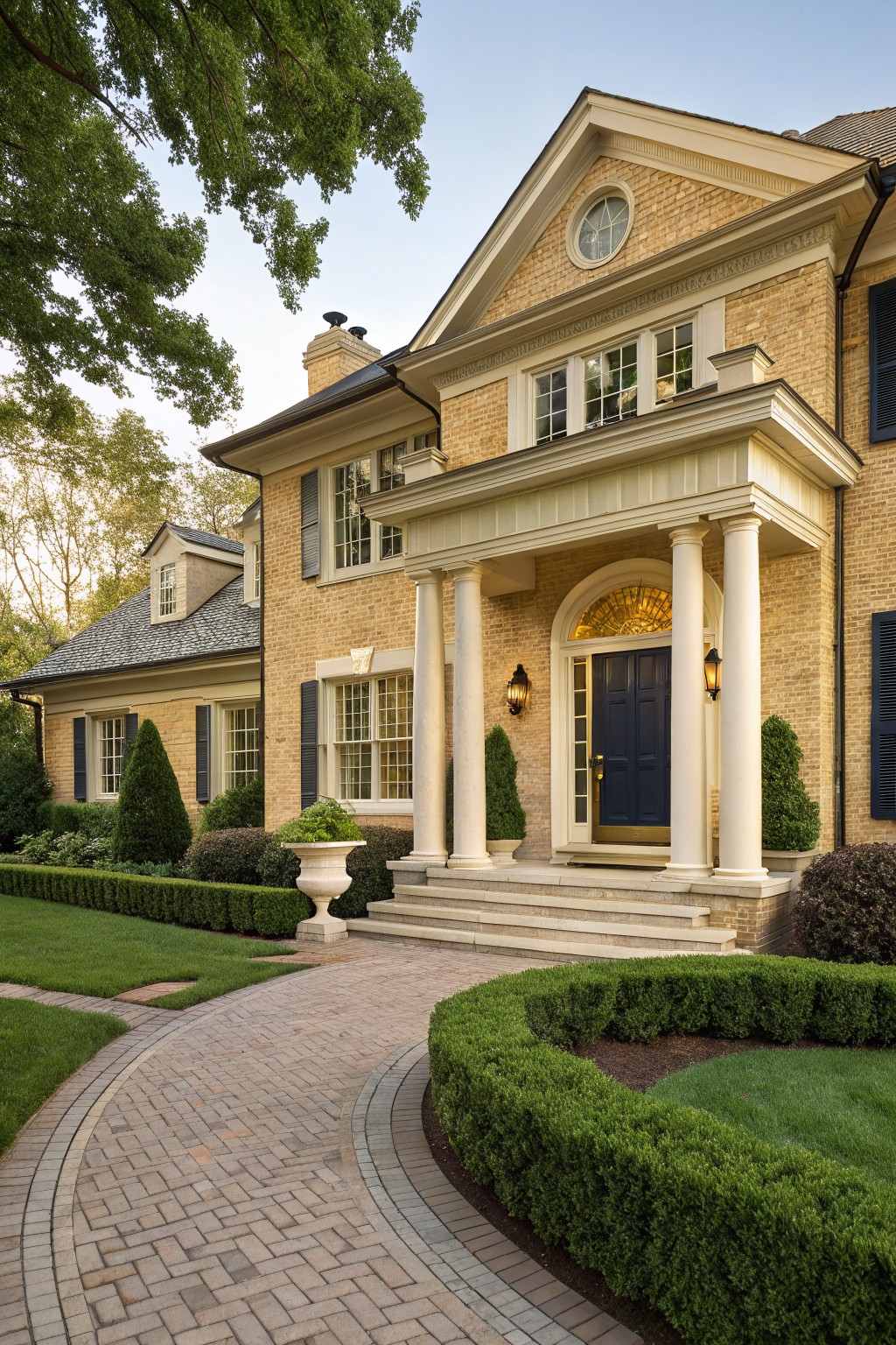 Two-story yellow brick house exterior with white columned portico, arched glass door in navy blue, flanked by lanterns and steps, surrounded by manicured shrubs and curved brick pathway.