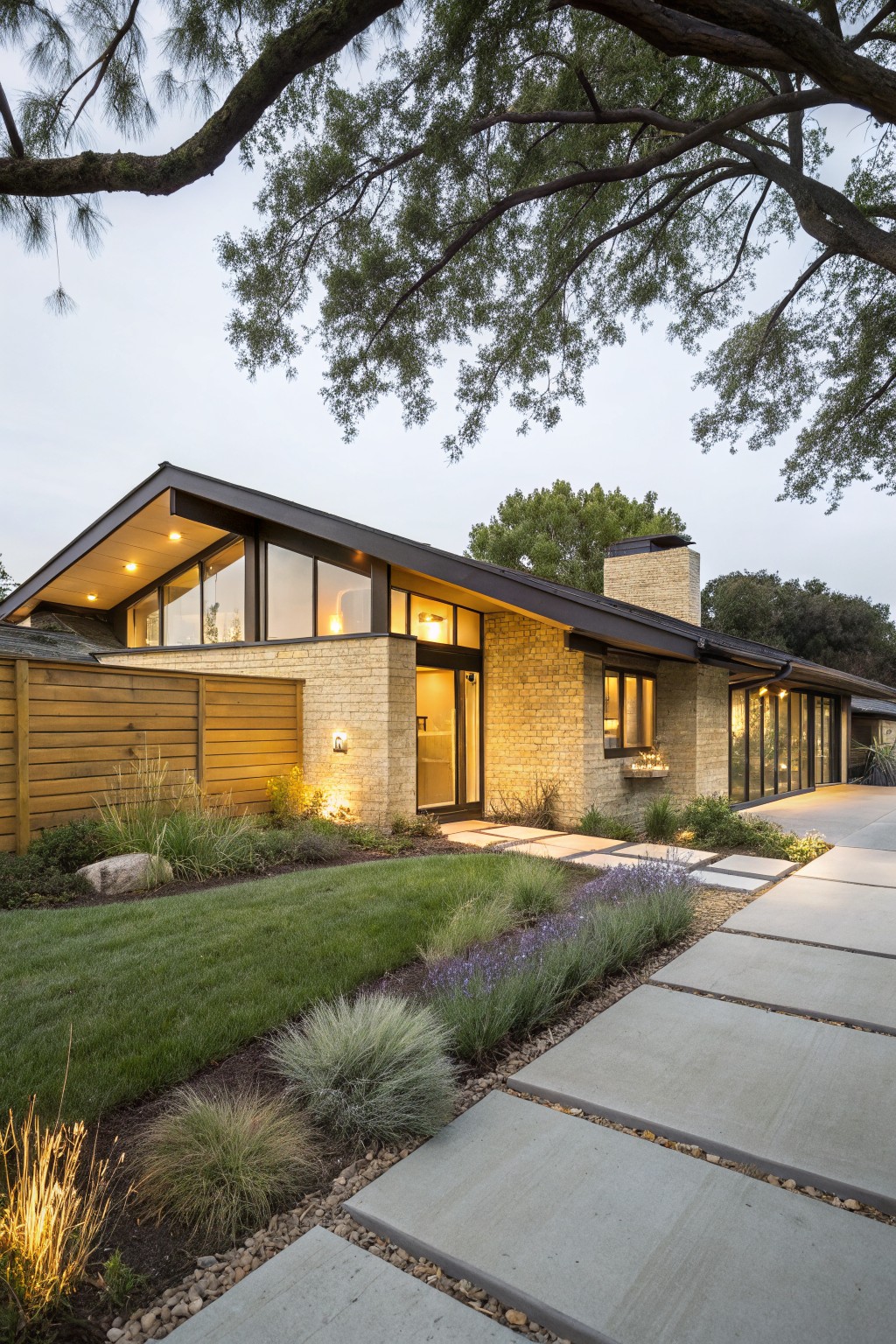 Single-story house exterior with light yellow textured brick walls, black metal-framed windows and sloped roof, glass entry door, brick chimney, wood fence, landscaped yard with grasses and pathway leading to the front.
