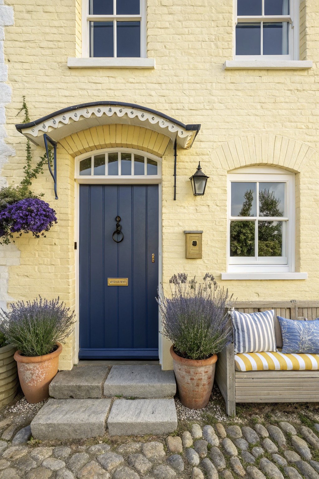 Pale yellow brick house exterior with navy blue front door under arched porch, white windows, lantern light, lavender plants in terracotta pots, wooden bench with cushions, and stone steps on cobblestone path.
