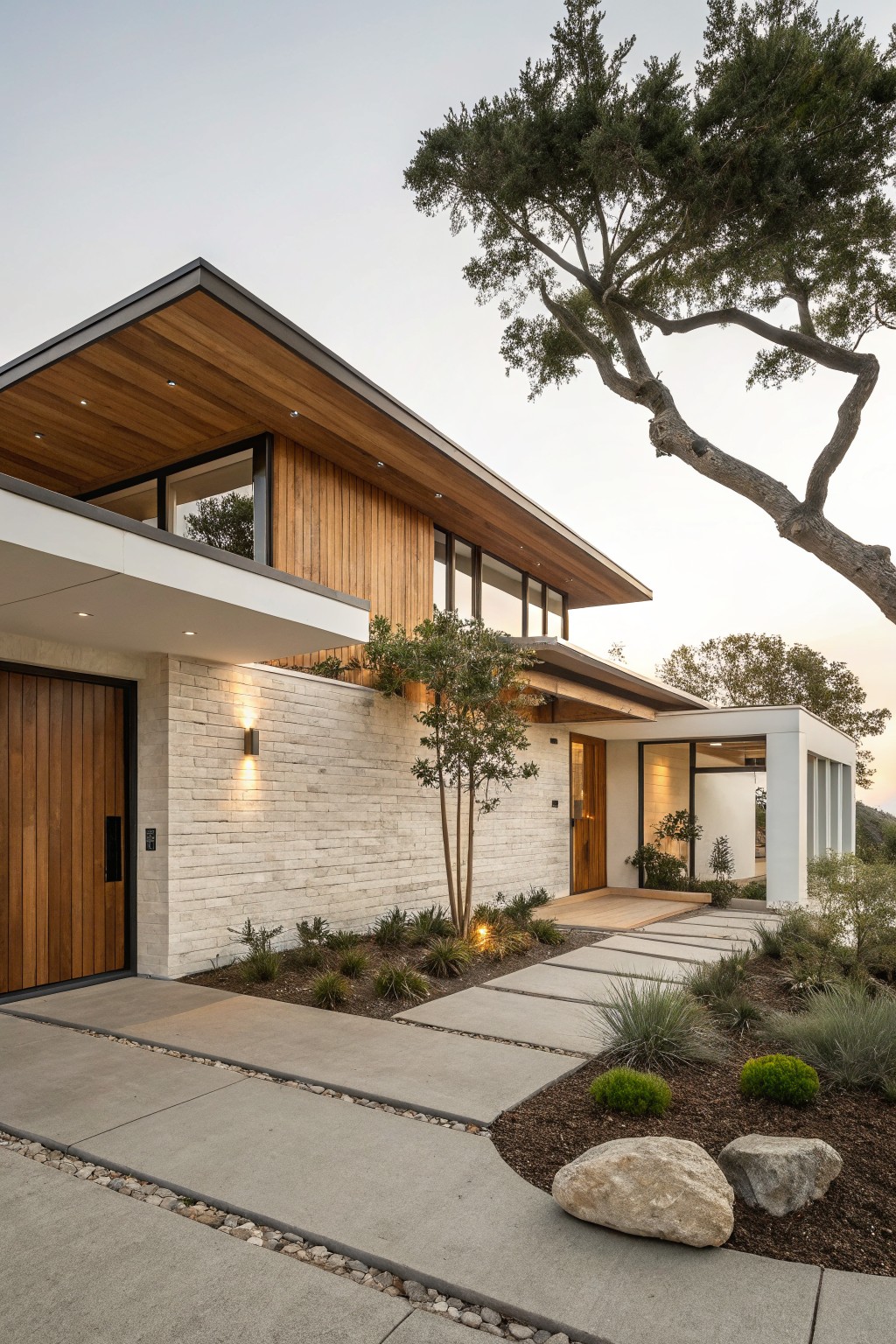 Contemporary two-story house exterior with white brick lower walls, vertical and horizontal wood cladding on upper sections and soffits, large pivoting wood entry door, lit pathway of concrete pavers, native shrubs, trees, and boulders in the front yard at dusk.
