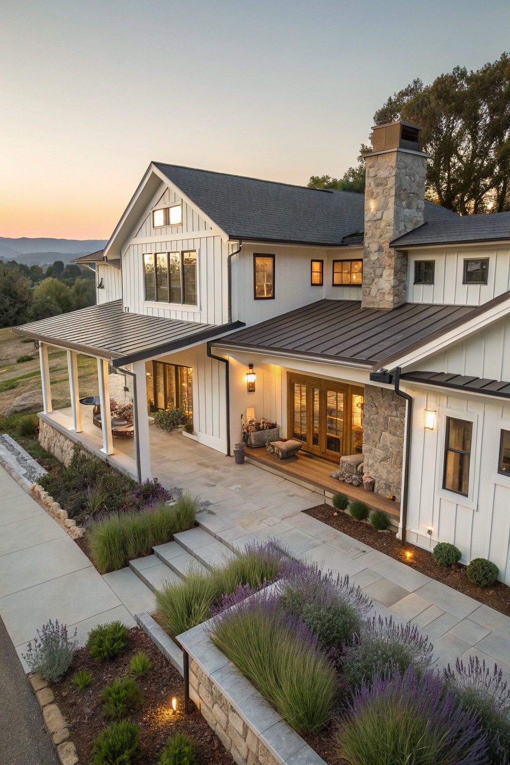 Aerial view of a two-story white board-and-batten house with dark standing-seam metal roof, stone chimney, covered porch supported by wood posts, stone entry steps, and drought-tolerant landscaping with lavender and grasses.
