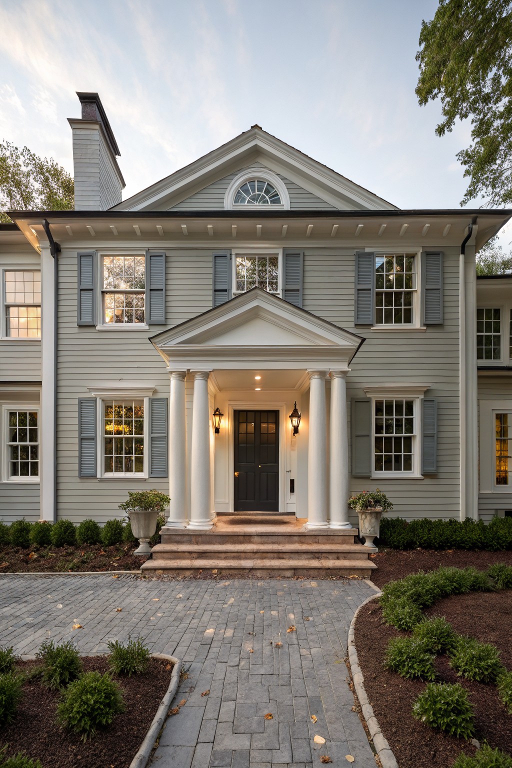 Front exterior of a light gray shingled house featuring a white-columned portico over a dark wood door, flanked by multi-pane windows with dark blue shutters, brick steps leading to a paver pathway, and low boxwood shrubs.