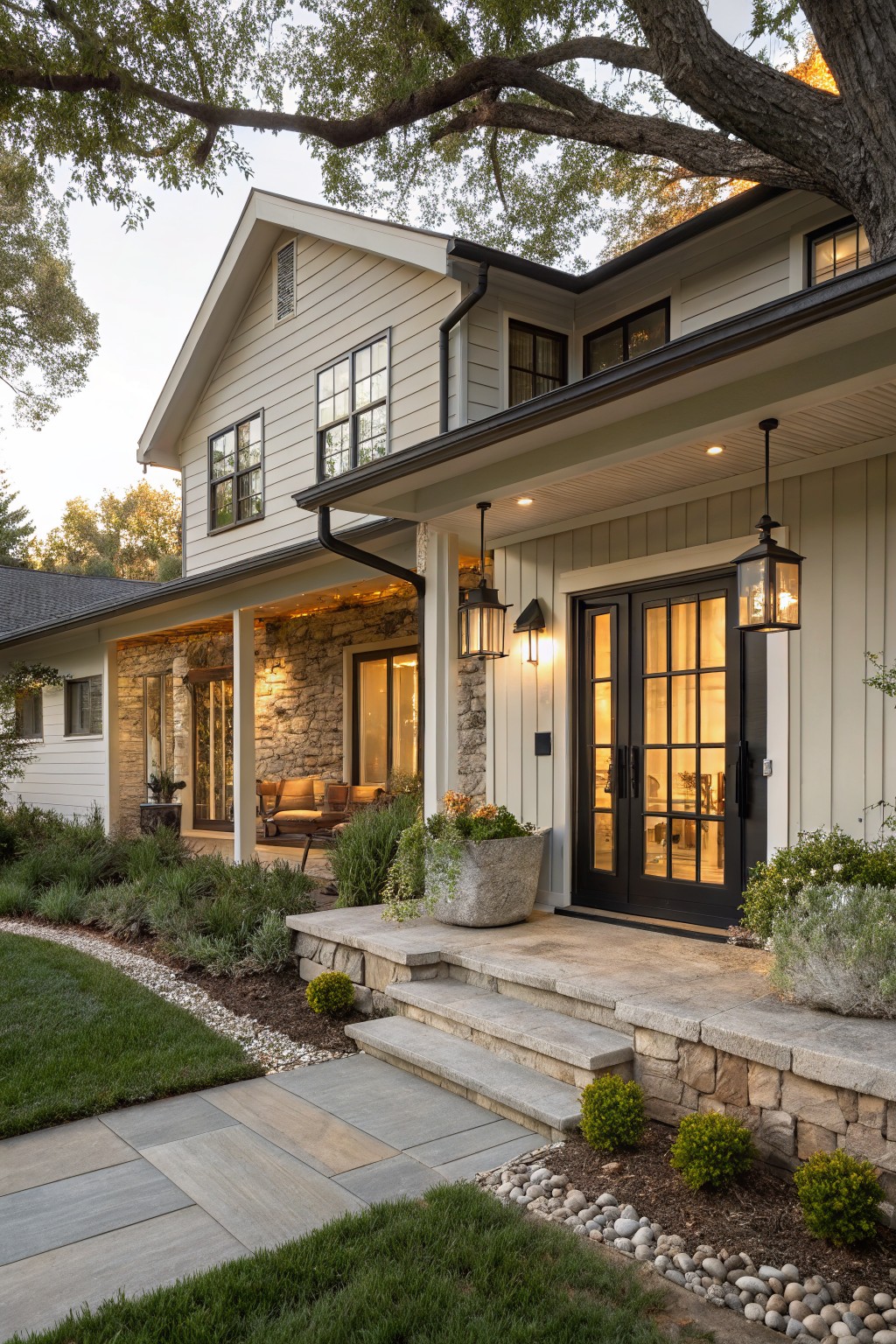 Two-story house exterior in light gray clapboard siding with covered front porch, black-framed double doors, stone base and steps, hanging lanterns, and landscaped yard with plants and gravel paths.